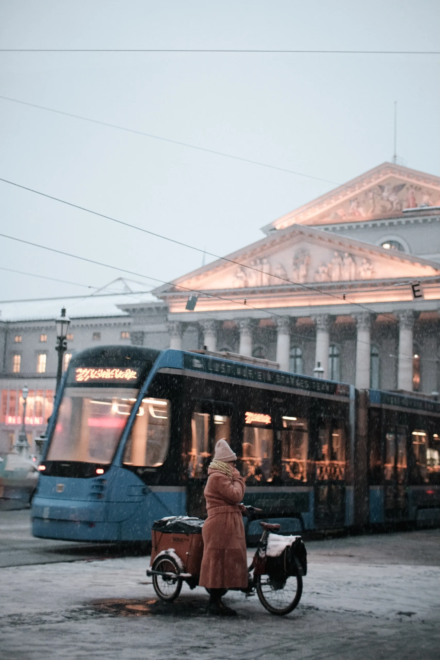 A tram in front of a columned building in winter, a woman with a cargo bike beside it.